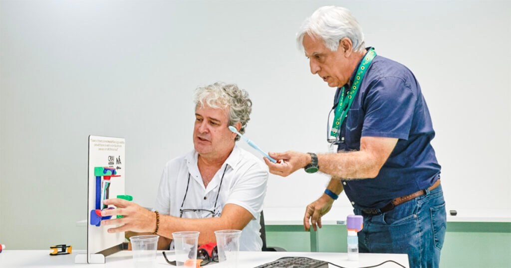 Duas pessoas em sala de aula manipulam um modelo científico colorido sobre a mesa, com copos plásticos e ferramentas ao redor.