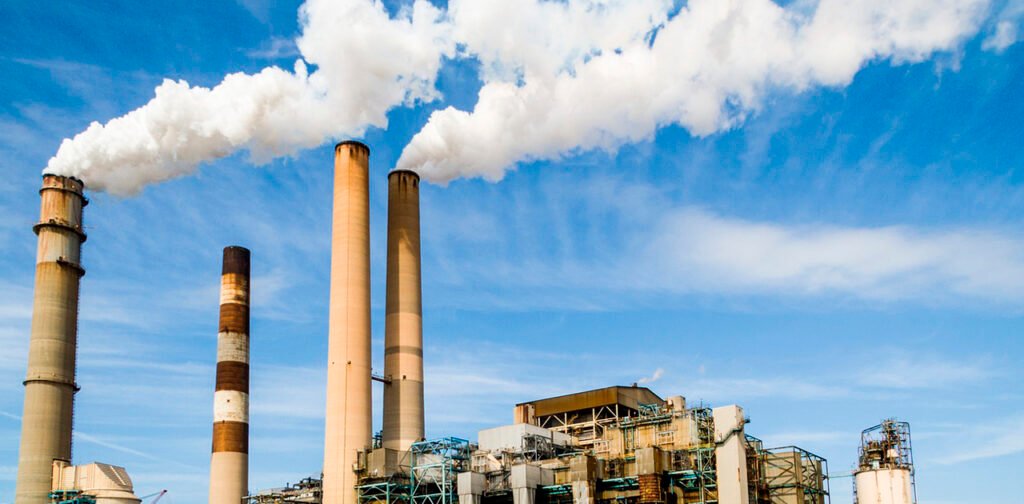 imagem é uma fotografia de uma usina termelétrica em funcionamento com quatro torres, sendo que em duas, saem fumaça branca