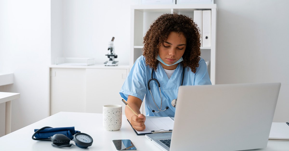 Mulher negra vestida com roupa azul, de uso hospitalar, escreve sobre uma mesa em que há um notebook, um copo e um aparelho de aferir pressão