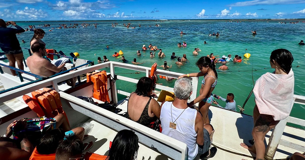 Imagem em plano aberto de uma paisagem marítima, com muitas pessoas nadando e outras em pé no que aparece um barco ou uma plataforma