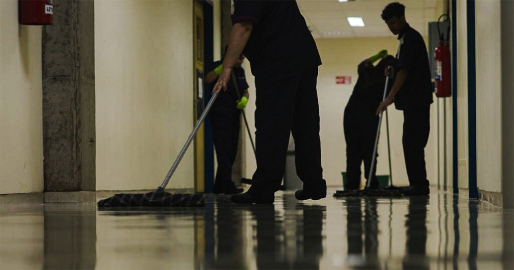 Quatro pessoas, de uniforme azul e luvas verdes, limpam o chão com rodos e panos.