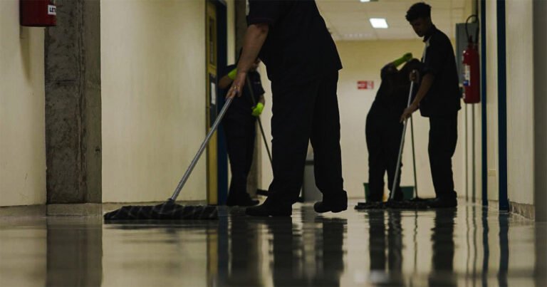Quatro pessoas, de uniforme azul e luvas verdes, limpam o chão com rodos e panos.