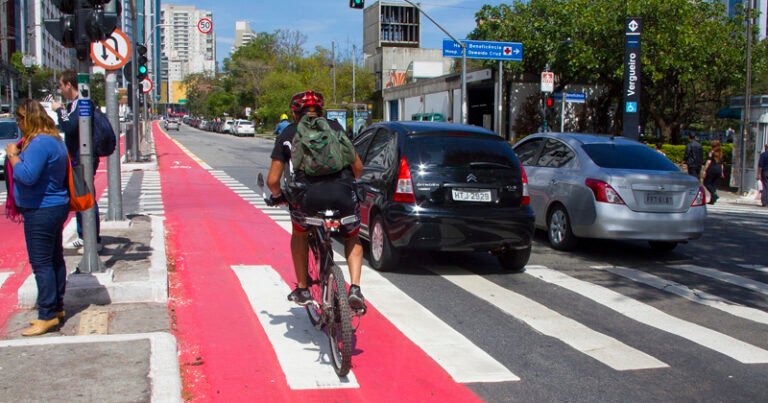 A imagem mostra uma cena urbana com uma ciclovia vermelha à esquerda e uma faixa de pedestres à direita. Um ciclista usando capacete e mochila está pedalando na ciclovia. Ao lado, há duas pessoas paradas na calçada, uma mulher de cabelo castanho e um homem de terno. Na rua, há dois carros parados próximos à faixa de pedestres. Ao fundo, é possível ver prédios e árvores, além de algumas placas de sinalização.