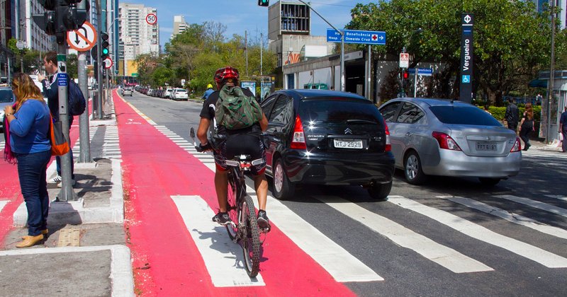 A imagem mostra uma cena urbana com uma ciclovia vermelha à esquerda e uma faixa de pedestres à direita. Um ciclista usando capacete e mochila está pedalando na ciclovia. Ao lado, há duas pessoas paradas na calçada, uma mulher de cabelo castanho e um homem de terno. Na rua, há dois carros parados próximos à faixa de pedestres. Ao fundo, é possível ver prédios e árvores, além de algumas placas de sinalização.