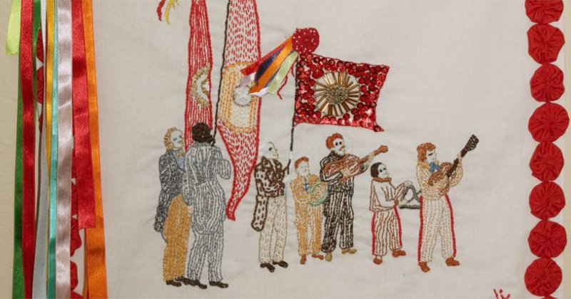 Detalhe de um bordado com pessoas numa festa e algumas delas segurando bandeira, ao lado tem algumas fitas