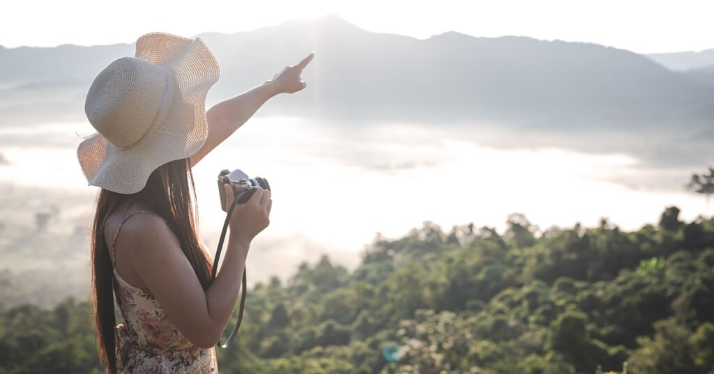 Imagem de uma jovem vestindo chapéu apontando com a mão esquerda um ponto no horizonte, enquanto com a mão direita segura uma máquina fotografica em meio a uma paisagem florestal