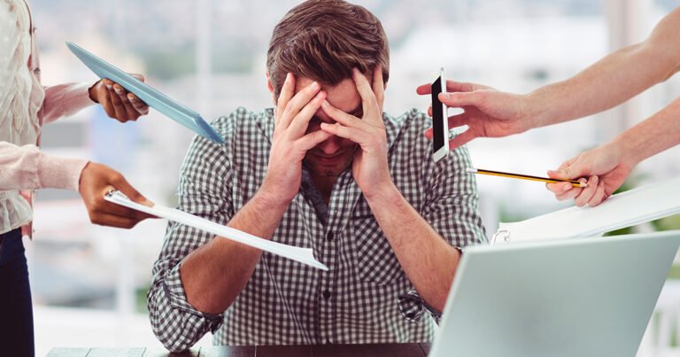 Homem jovem, de camisa xadrez, está sentado à mesa com as mãos na cabeça, demonstrando estresse ou sobrecarga. Ao seu redor, mãos de outras pessoas estendem objetos como papéis, um tablet, um celular e um lápis em sua direção.