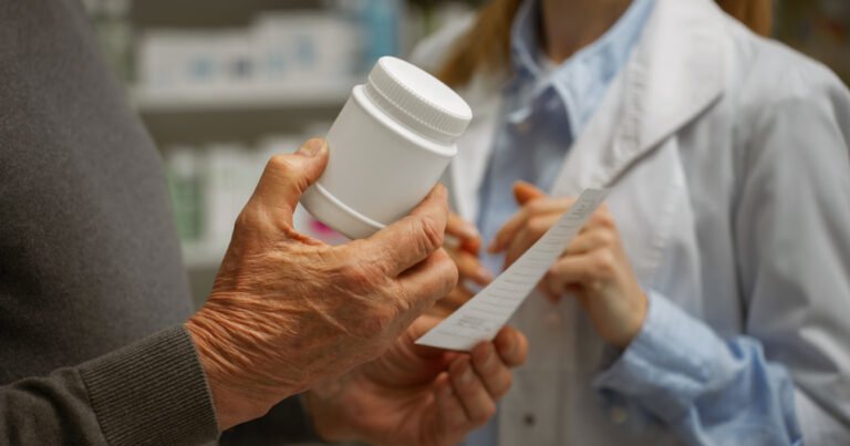 Foco nas mãos de duas pessoas, uma segurando um pote de medicamentos e um papel com a receita médica e outra ao lado de jaleco branco gesticulando