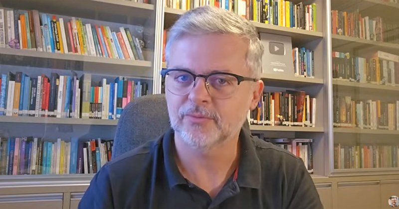 Homem branco com barba, bigode e cabelos grisalhos sentado numa cadeira e ao fundo uma estante cheia de livros