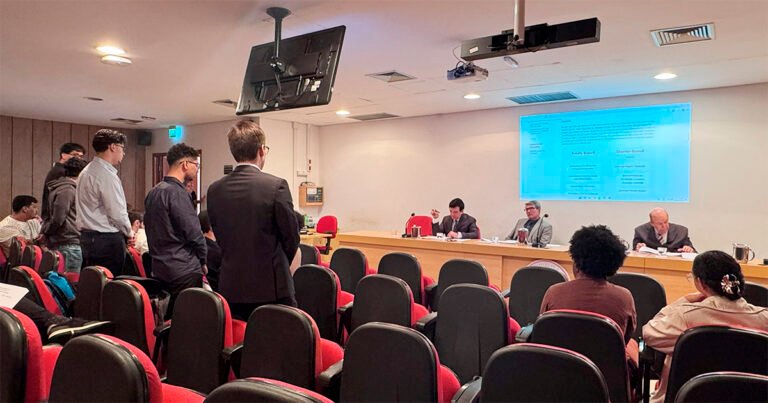 Auditório com três homens sentados atrás de uma mesa grande e várias pessoas em pé e sentadas nas cadeiras vermelhas, diante da mesa, participando de um julgamento.