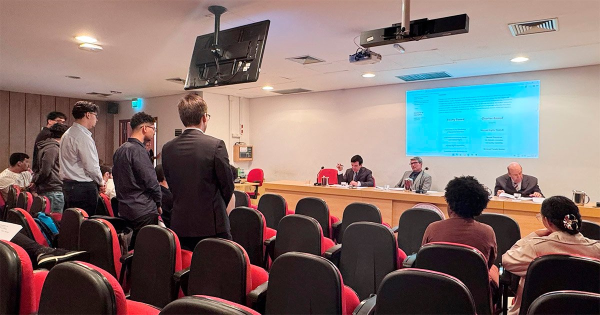 Auditório com três homens sentados atrás de uma mesa grande e várias pessoas em pé e sentadas nas cadeiras vermelhas, diante da mesa, participando de um julgamento.