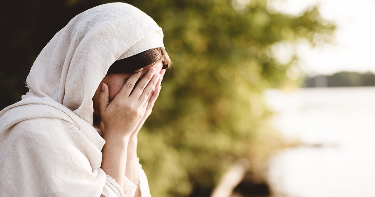 A imagem mostra uma mulher usando um véu claro, com o rosto coberto pelas mãos, em um ambiente externo com natureza ao fundo.
