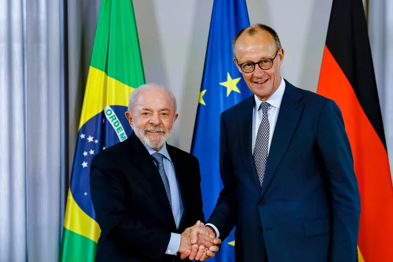 O presidente Lula (esquerda) aperta a mão do chanceler da Alemanha, Friedrich Merz. Ao fundo está a bandeira brasileira, a da União Europeia e a da Alemanha