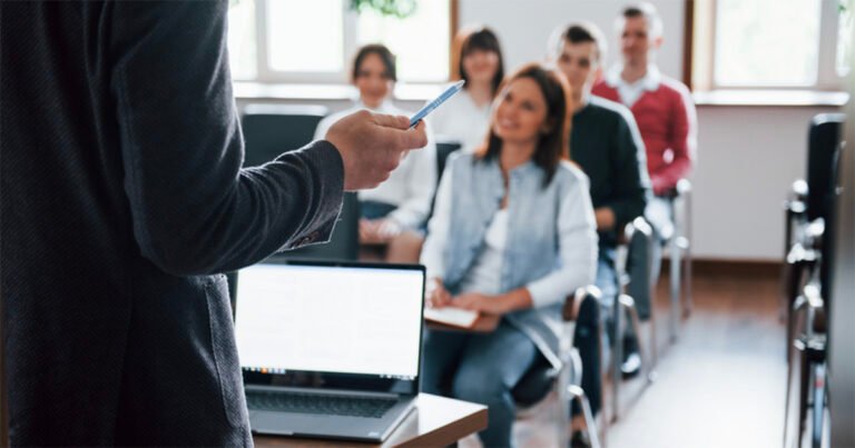 Imagem de uma mão direita branca, segurando uma caneta e explicando algo a uma platei em sala de aula