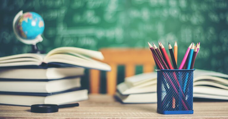 Imagem de uma mesa escolar contendo material de estudo, como lápis e livros empilhados uns sobre os outros, além de um pequeno mapa mundi