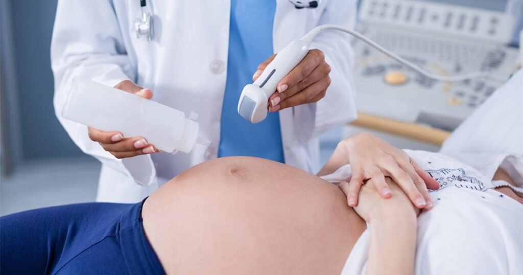 Médica realizando um ultrassom numa gestante com gravidez avançada. A gestante utiliza calça azul e blusa branca e está com a barriga aparente e as mãos cruzadas acima, e a médica usa jaleco branco e estetoscópio no pescoço e segura o aparelho de ultrassom.