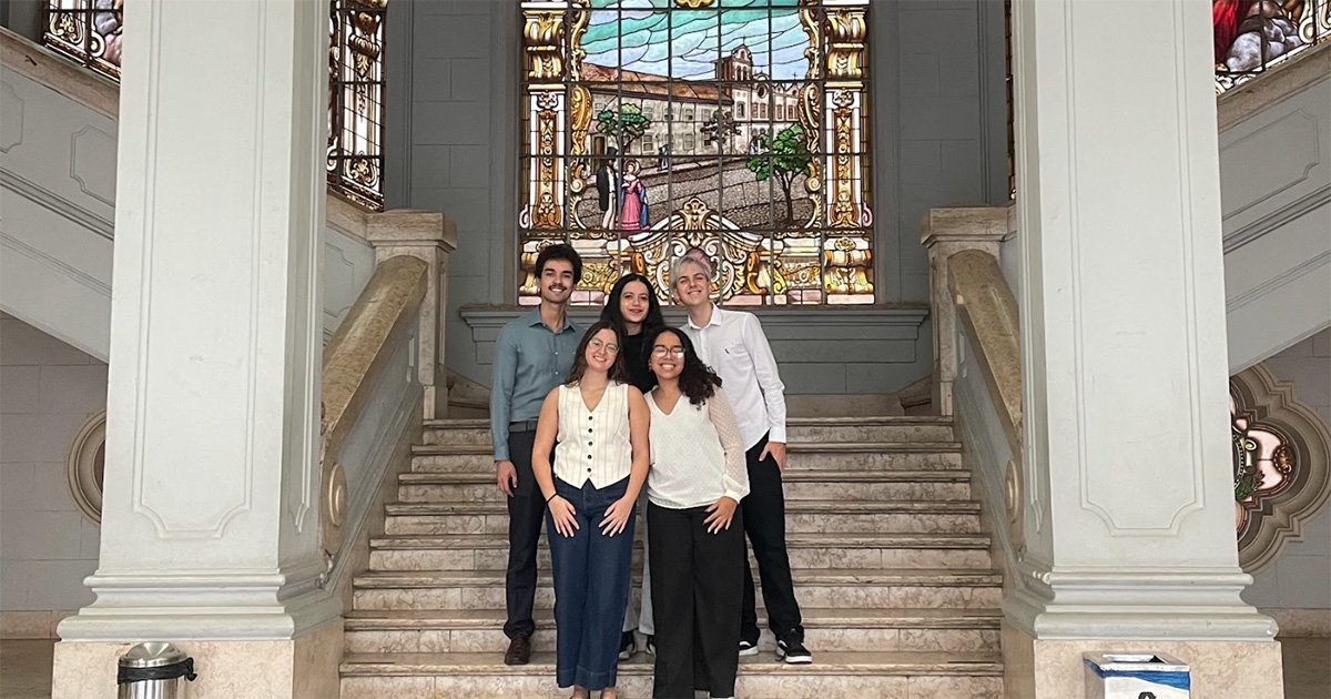 Dois homens e três mulheres de roupas sociais sorriem para foto em escadaria, com vitral ao fundo.