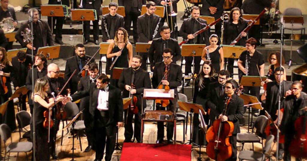 Membros de uma orquestra em pé, num palco.