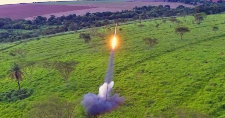 Área aberta com gramado e árvores ao fundo e no centro um foguete em lançamento com fumaça e fogo saindo da sua base