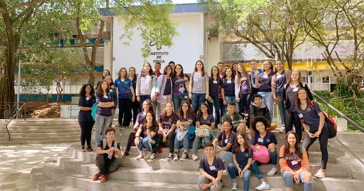 Grupo de estudantes, alguns em pé e outros sentados nos degraus de uma escadaria, em frente a um prédio baixo