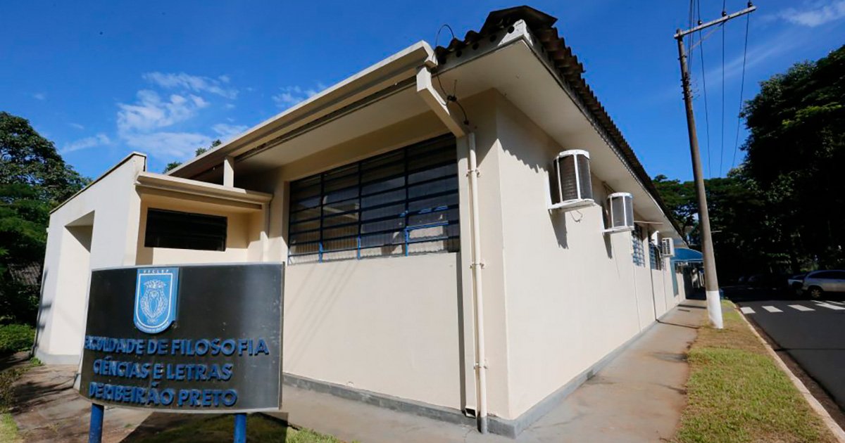 A imagem mostra um prédio com paredes claras e telhado inclinado. Na parte frontal, há um letreiro azul com o nome "Faculdade de Filosofia, Ciências e Letras de Ribeirão Preto" e um brasão. O edifício tem janelas com grades e várias unidades de ar-condicionado instaladas. Há uma rua ao lado com postes de energia elétrica, e o céu está azul com algumas árvores ao fundo