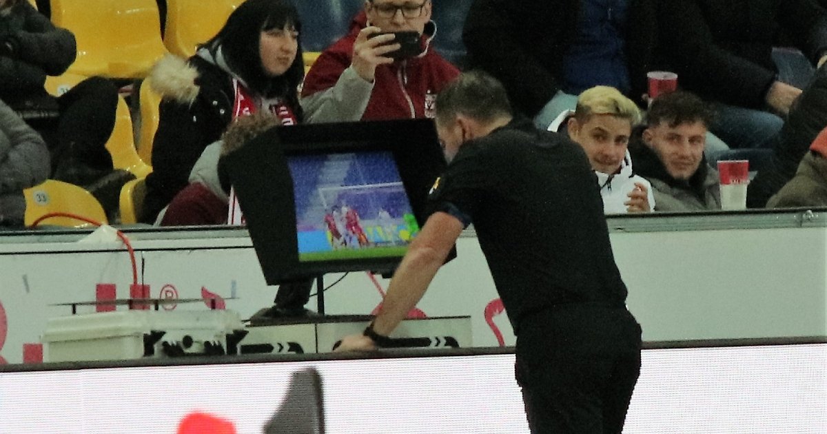 Árbitro de futebol com uniforme preto, visto de costas, consulta o monitor de vídeo do VAR que mostra um lance da partida, enquanto é observado por torcedores