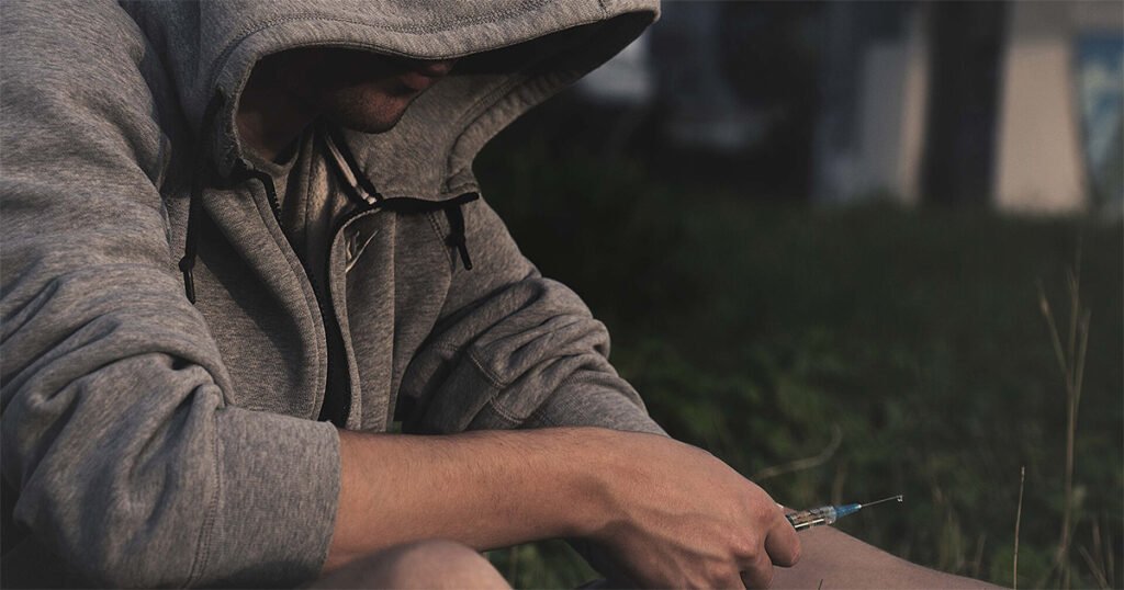 Jovem sentado em um gramado, usando blusa de capuz que encobre o rosto, segurando uma seringa de injeção perto do braço