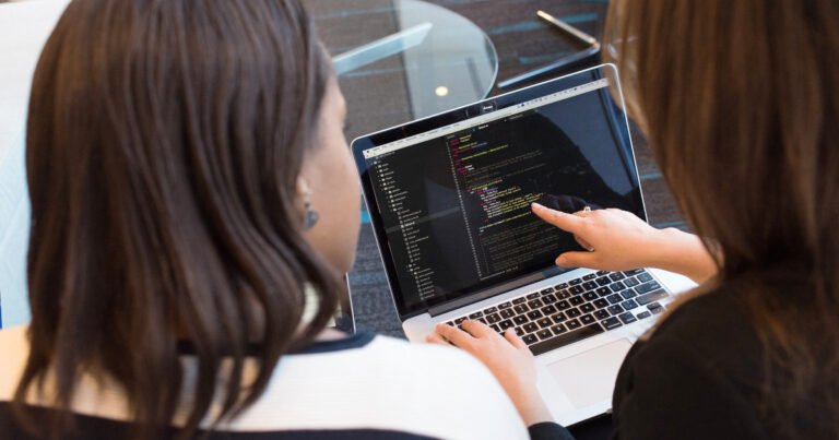 Duas mulheres analisam um código de programação exibida na tela de um notebook