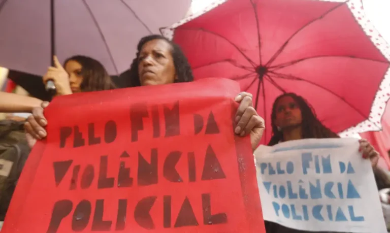 Manifestantes realizam protesto contra a violência policial. Mulher segura cartaz feito em cartolina vermelha onde se lê: Pelo fim da violência policial
