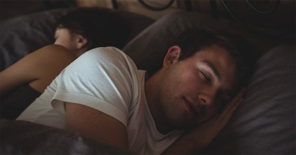 Homem dorme de lado em uma cama, usando camiseta branca, com o rosto apoiado sobre um travesseiro cinza