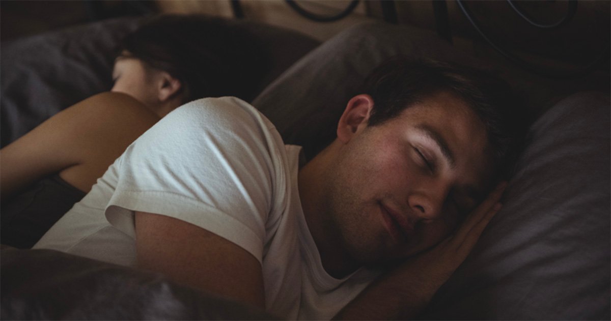 Homem dorme de lado em uma cama, usando camiseta branca, com o rosto apoiado sobre um travesseiro cinza