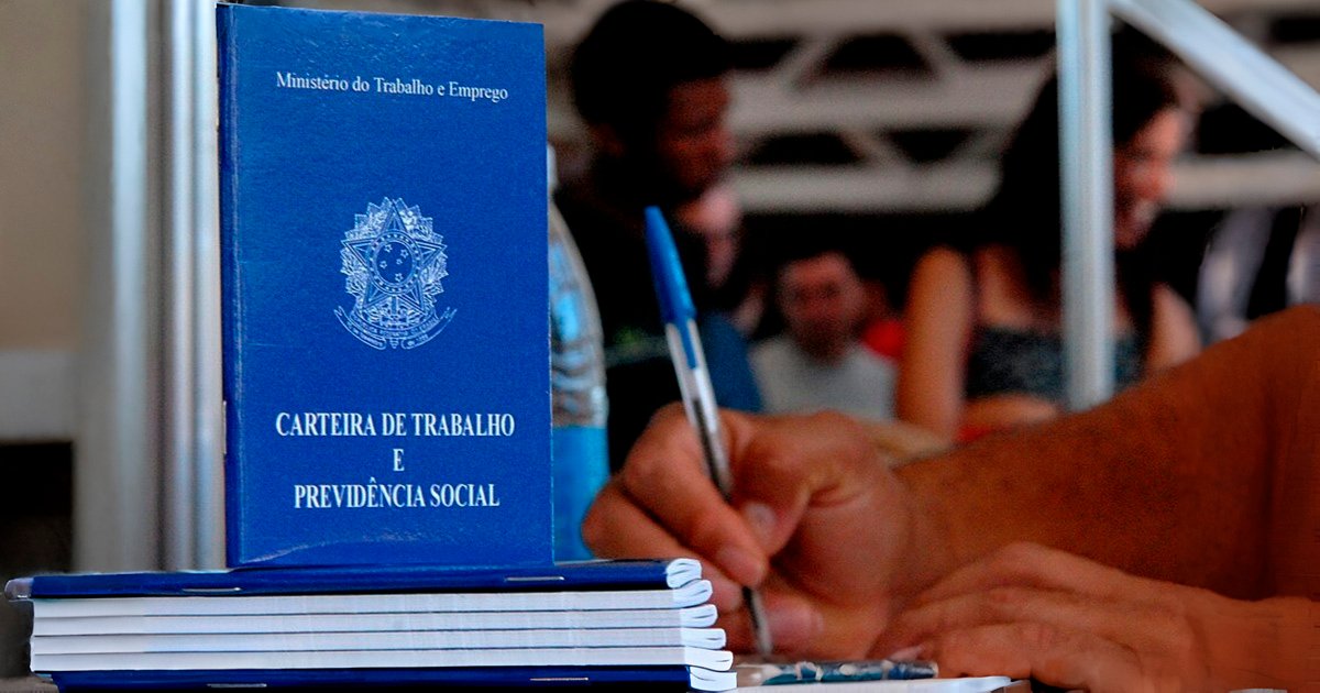 Fotomontagem mostrando em primeiro plano uma carteira de trabalho e uma mão empunhando uma caneta e assinando um documento; ao fundo, desfocadas, aparecem várias pessoas