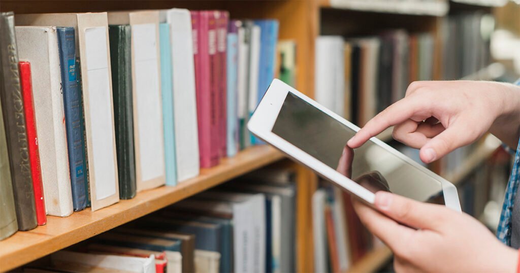 Uma estante com livros em um biblioteca, no canto direito, em frente à estante, uma pessoa usa um tablet.