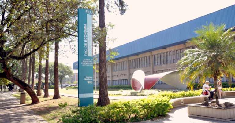 Prédio de arquitetura moderna cercado por árvores, jardins e áreas de circulação. Em frente, há um gramado, uma escultura e pessoas sentadas e caminhando pelo local