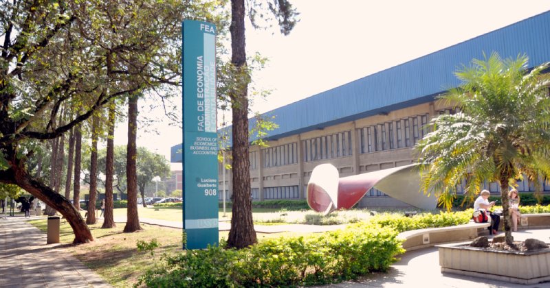 Prédio de arquitetura moderna cercado por árvores, jardins e áreas de circulação. Em frente, há um gramado, uma escultura e pessoas sentadas e caminhando pelo local