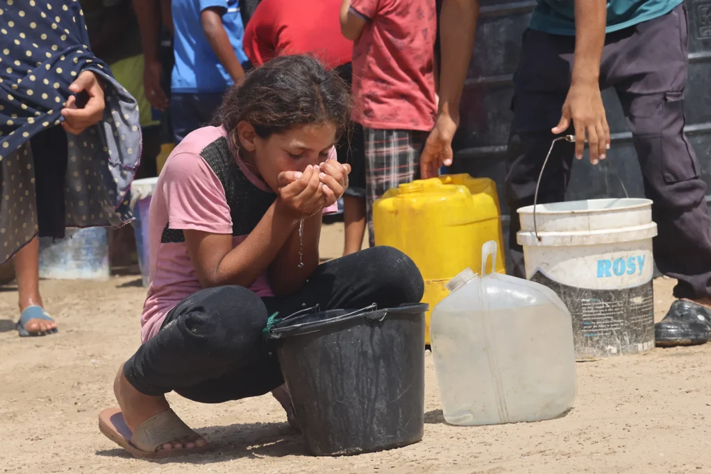 Uma menina bebe água de um balde de plástico enquanto palestinos deslocados coletam água na área de Mawasi, em Khan Yunis, em 14 de agosto de 2025. À época, organizações internacionais já trabalhavam para fornecer água potável à população em meio ao agravamento da crise e à disseminação de doenças devido à destruição do sistema de saneamento básico, causada por Israel.