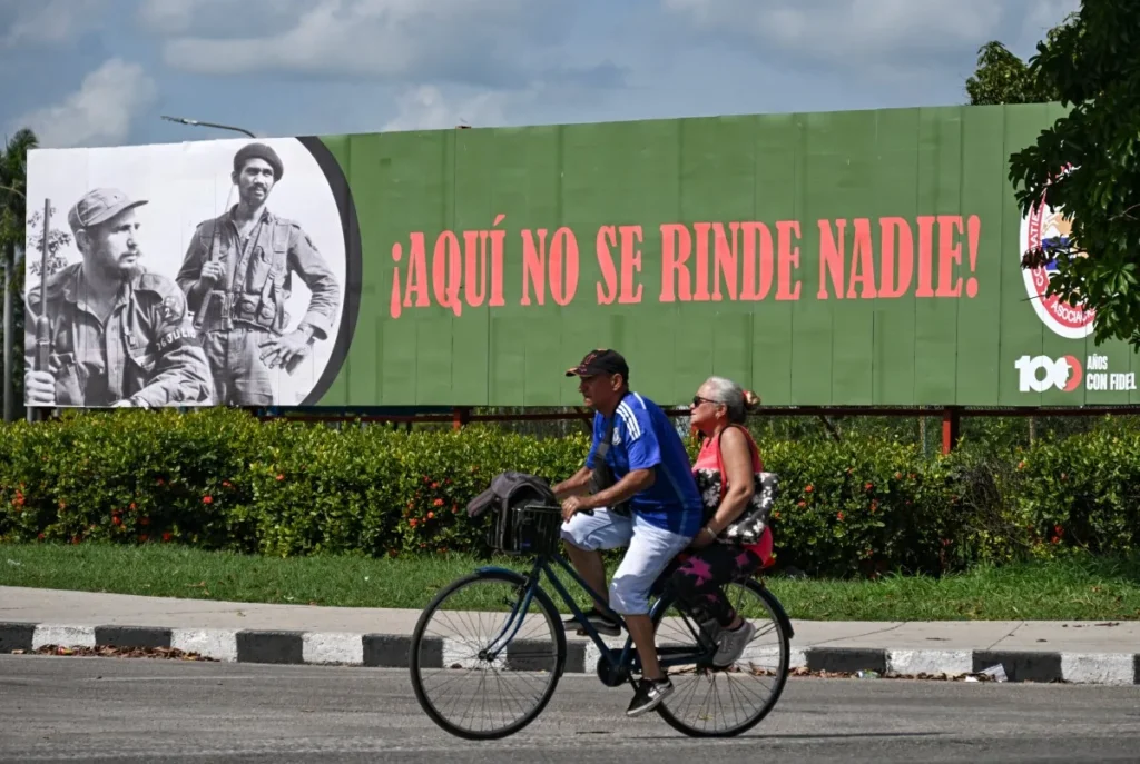 Um homem e uma mulher andam de bicicleta em frente a uma placa com a imagem do líder revolucionário cubano Fidel Castro (1926-2016) e do comandante Juan Almeida Bosque (1927-2009), com a inscrição "Ninguém se rende aqui", em Havana, em 19 de junho de 2026.