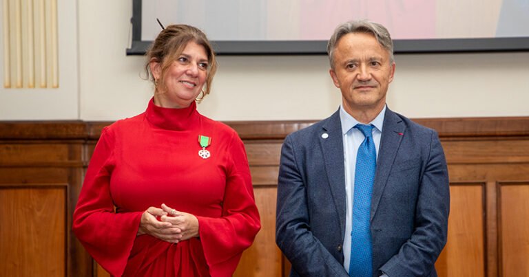 Mulher branca de cabelos loiros escuros presos atrás da cabeça e de vestido vermelho ao lado de homem branco de cabelos grisalhos e terno com gravata azul